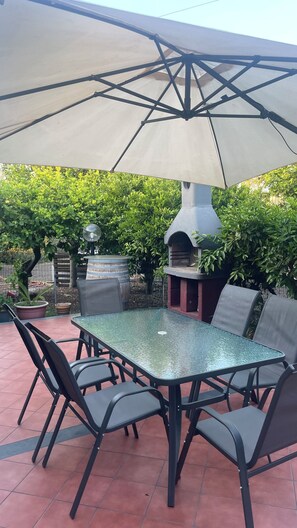 An outdoor patio with a glass-top table and chairs, a large umbrella, and a barbecue grill. - Cozy apartment with a view of Mount Etna, a spacious garden, and a relaxing atmosphere. No pets. (Aci Sant’Antonio)