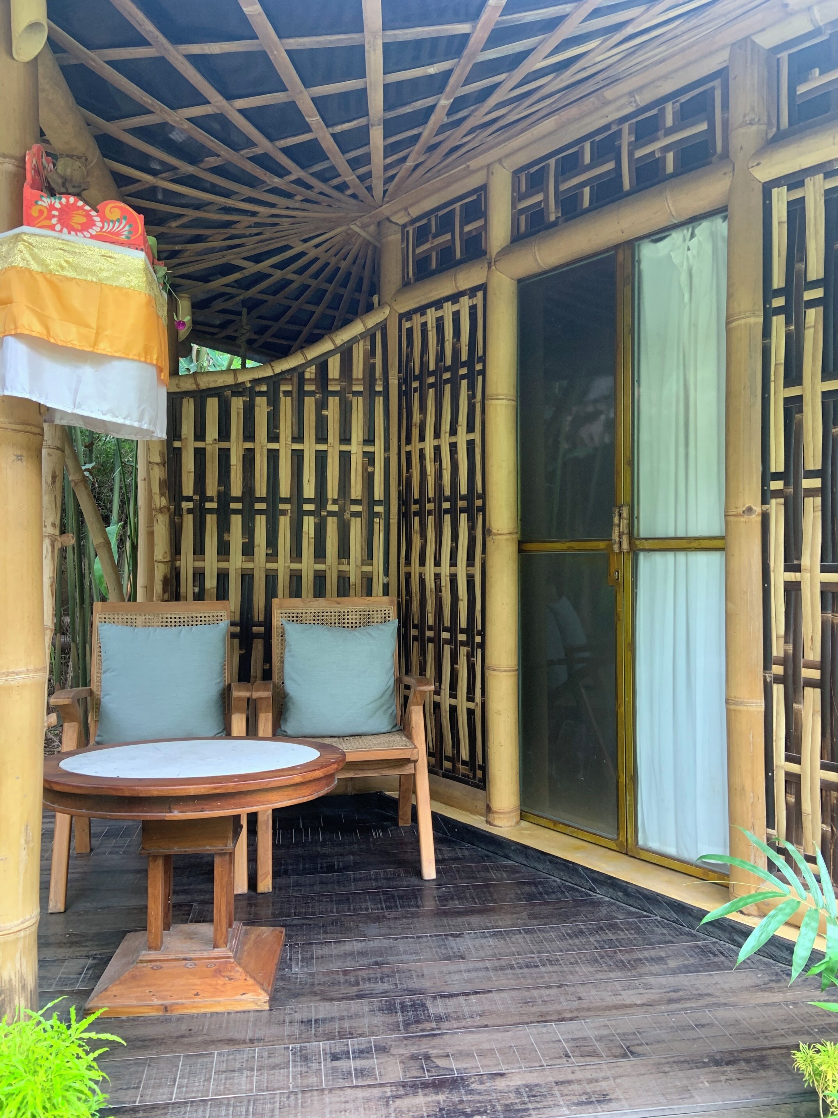 Un espacio de estar al aire libre cubierto, con dos sillas, una mesa, columnas de bambú y un amplio ventanal corredera.