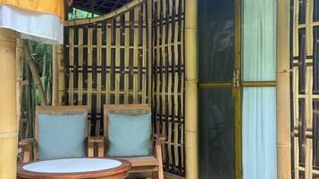 Un espacio de estar al aire libre cubierto, con dos sillas, una mesa, columnas de bambú y un amplio ventanal corredera.