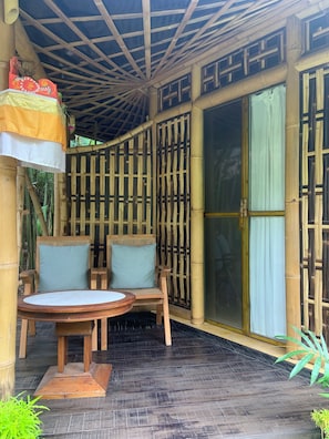 A covered outdoor seating area with two chairs and a table, bamboo columns, and a sliding glass door. - Swarma Villas (Gianyar)