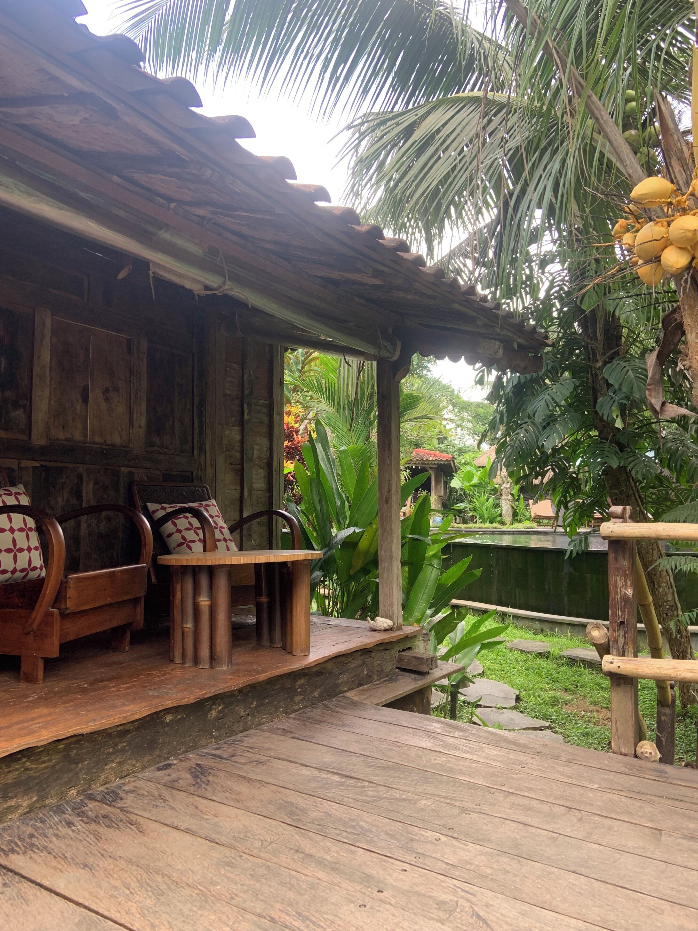 A wooden deck with a rustic outdoor seating area, including a bench and chairs, surrounded by lush greenery and a palm tree.