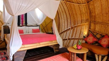 A room with a thatched roof, a bed with a canopy, a small round table, and a cushioned bench.
