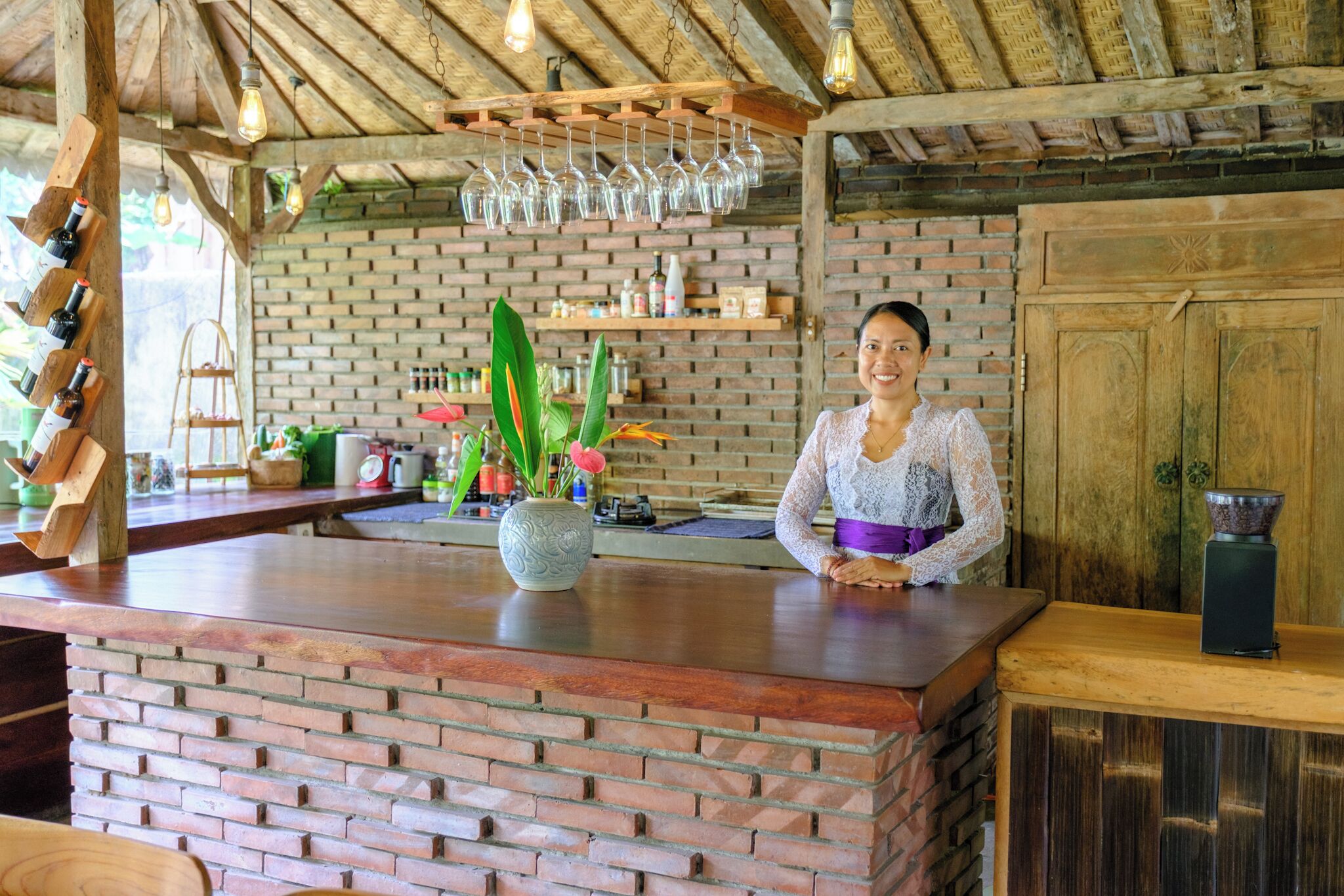 Zona de bar con mostrador de ladrillo, copas de vino colgantes y una variedad de botellas y plantas.