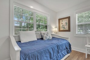 Een slaapkamer met een bed, witte wanden, een raam met zonwering, een nachtkastje met een lamp en een lijst met een afbeelding aan de muur.