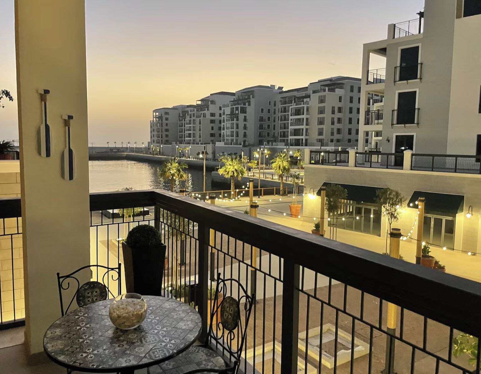 A balcony with a table and chairs, overlooking a waterfront with buildings and palm trees.