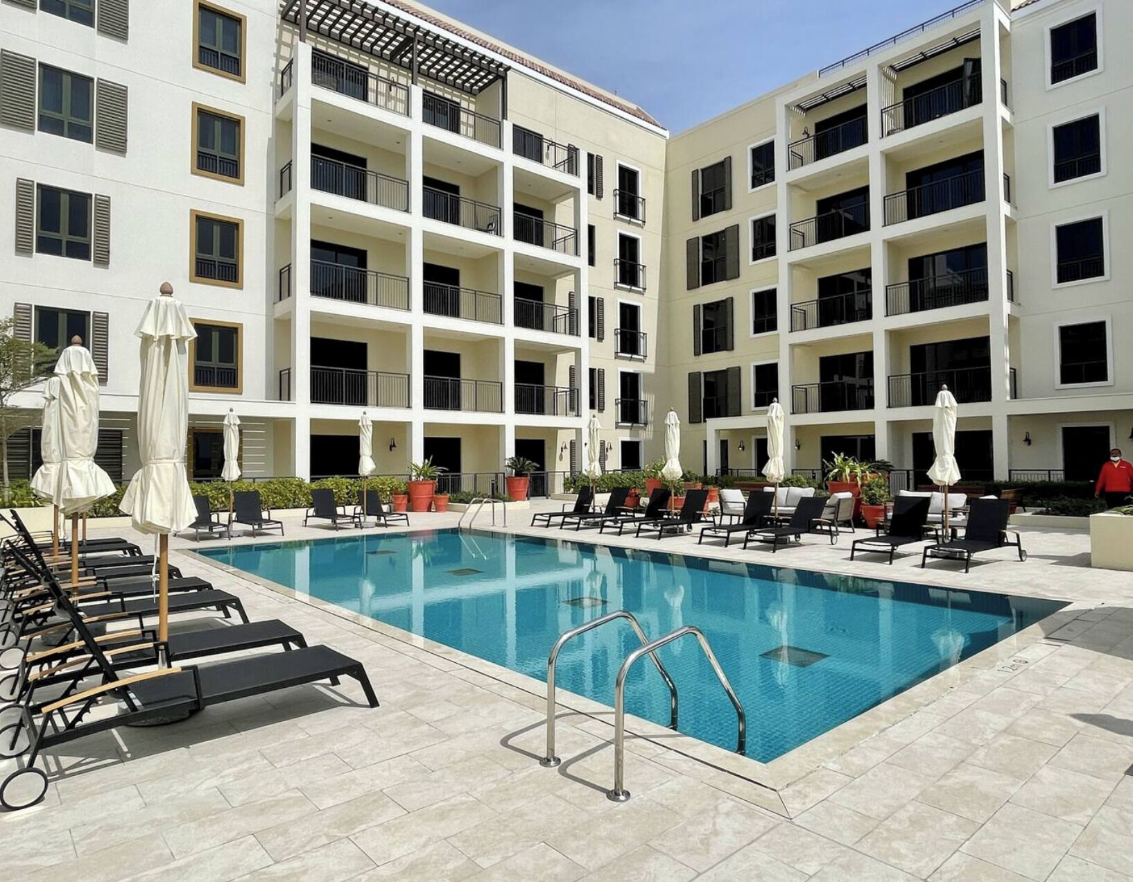 A hotel pool area with multiple lounge chairs and umbrellas, surrounded by a multi-story building with balconies.