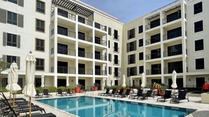 Zona de piscina de hotel con múltiples sillas de descanso y sombrillas, rodeada por un edificio de varios pisos con balcones.