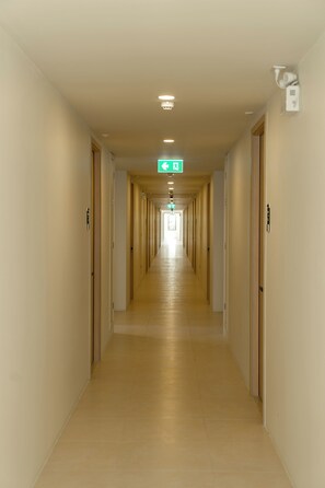 Un largo y estrecho pasillo de hotel con puertas a ambos lados, un letrero de salida de emergencia verde y un accesorio de iluminación en el techo.