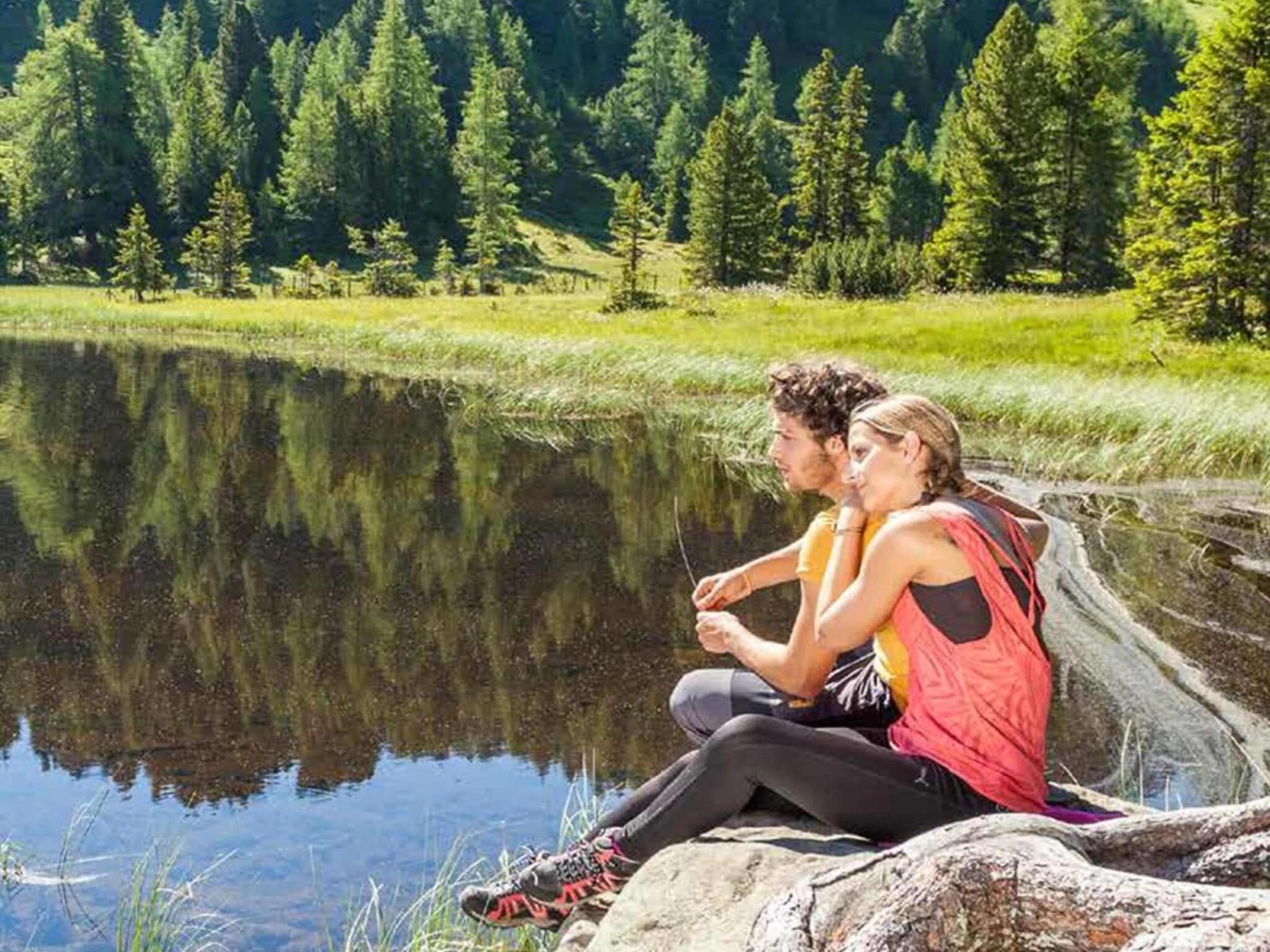 Dos personas sentadas junto a un lago tranquilo, con un bosque al fondo.