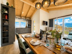 A dining area with a wooden table set for a meal, a kitchen in the background, and a view of the outdoors. - Nice ski-in-ski-out lodge with sauna and balcony (Sankt Lambrecht)