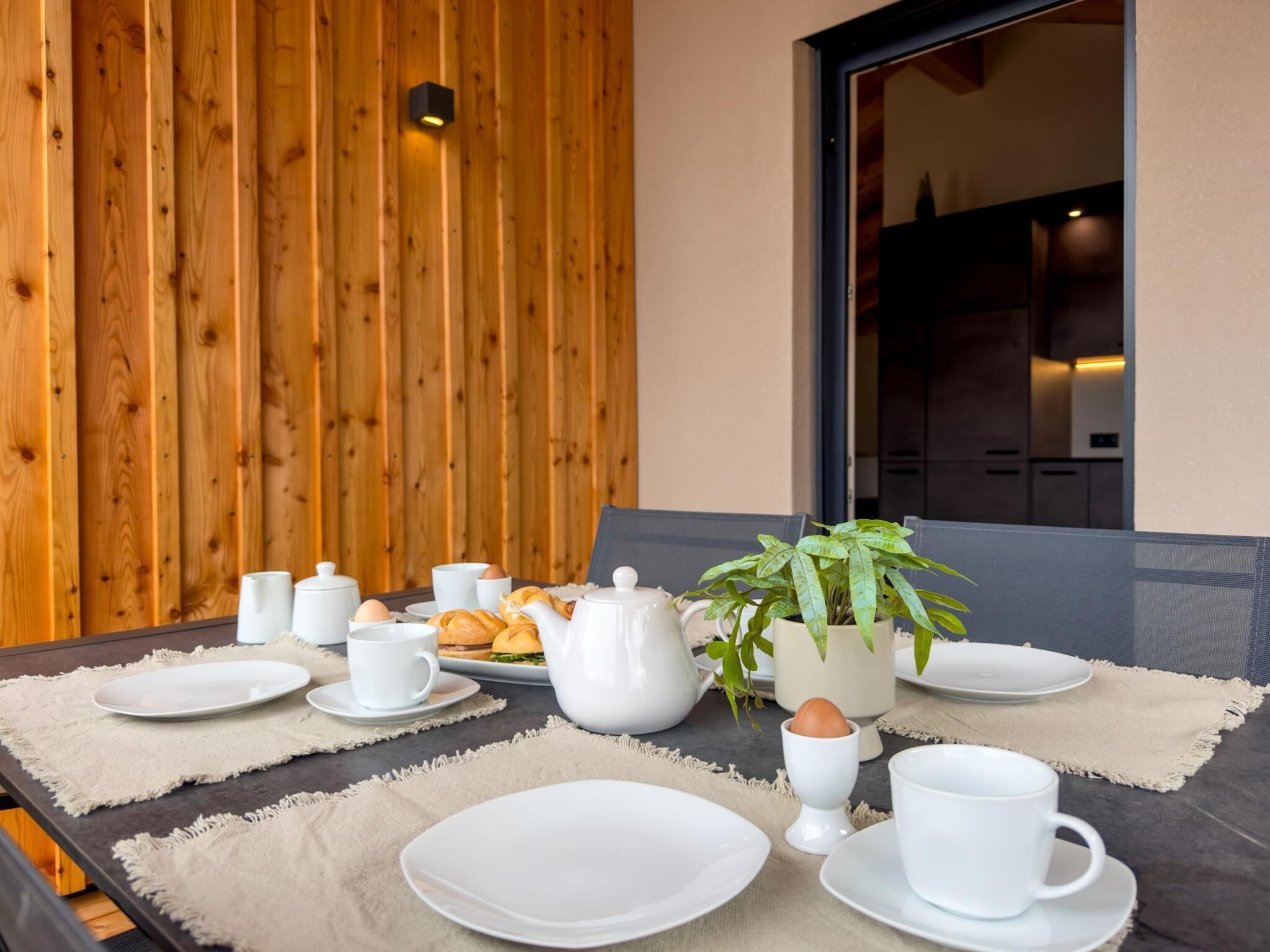 Una mesa de comedor dispuesta con platos blancos, tazas y una tetera, acompañada por una planta y una cesta de comida.