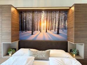 Une chambre à coucher avec un grand lit, une tête de lit et une œuvre d’art représentant un paysage forestier, accrochée au mur.