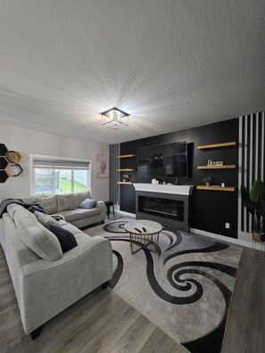 A modern living room with a sofa, a coffee table, a fireplace, a TV, and decorative shelves. - Treasure Villa: Prestigious Lodge | King Bed | AC (Edmonton)