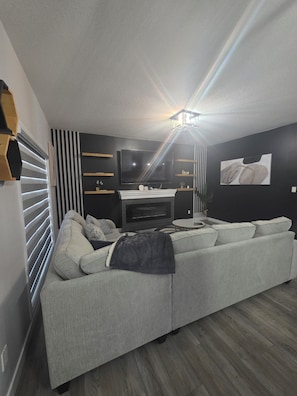 A modern living room with a grey sofa, a flat-screen TV, and a fireplace. - Treasure Villa: Prestigious Lodge | King Bed | AC (Edmonton)