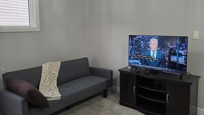 A living room with a grey sofa, a television on a black stand, and a ceiling light fixture. - Treasure Villa: Prestigious Lodge | King Bed | AC (Edmonton)