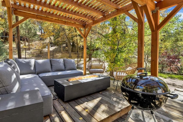 An outdoor patio with a wooden pergola, a gray sectional sofa, a round grill, and a wicker chair.