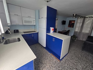 A compact kitchen with white and blue cabinets, a microwave, a refrigerator, a sink, and a small dining area with a table and chairs. - Puntilla Deluxe Apartment (Cataño)
