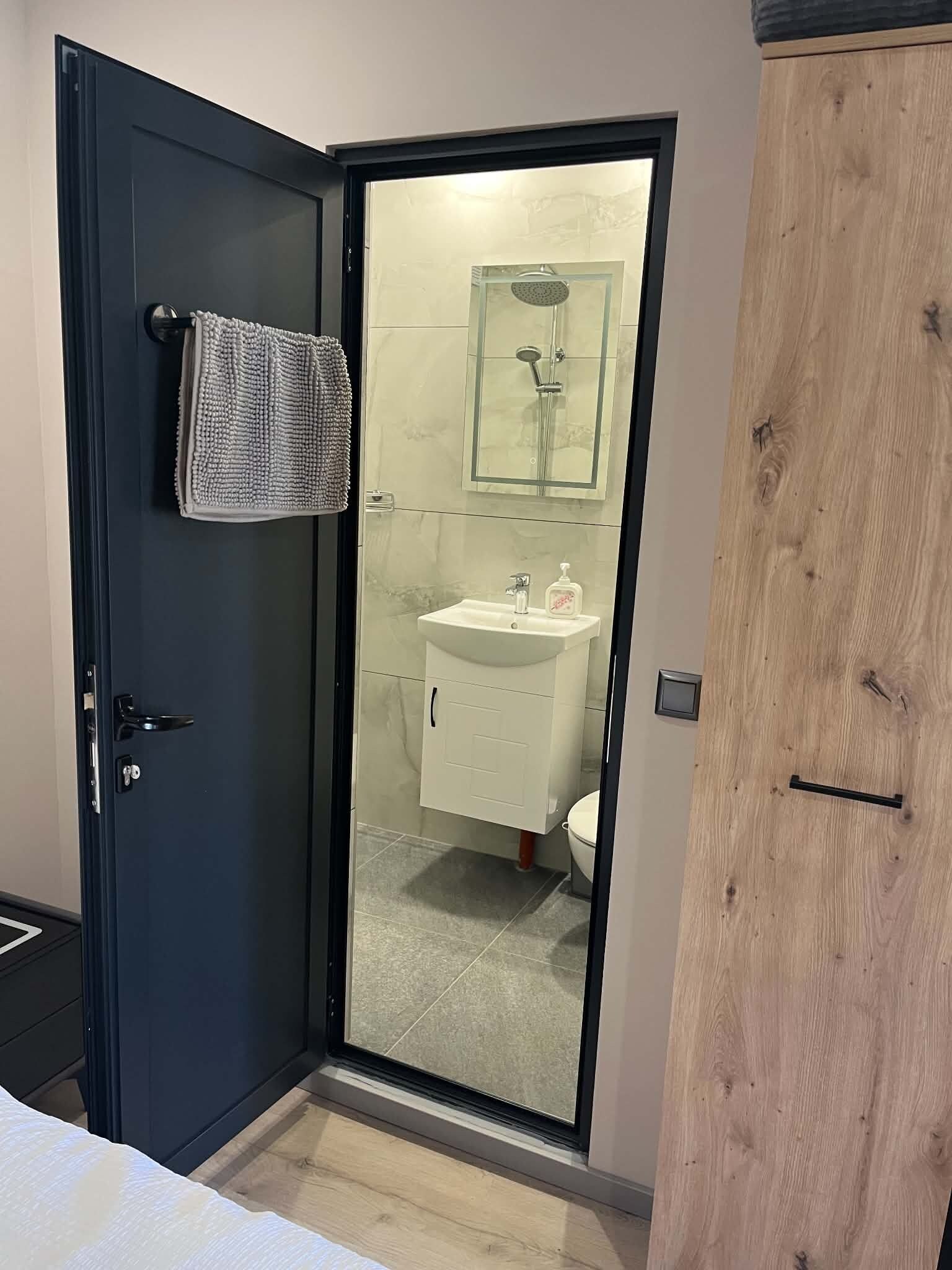 A room with a dark door, a towel hanging on the door, a glass shower enclosure, a white sink, and a wooden wardrobe.