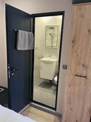 A room with a dark door, a towel hanging on the door, a glass shower enclosure, a white sink, and a wooden wardrobe.
