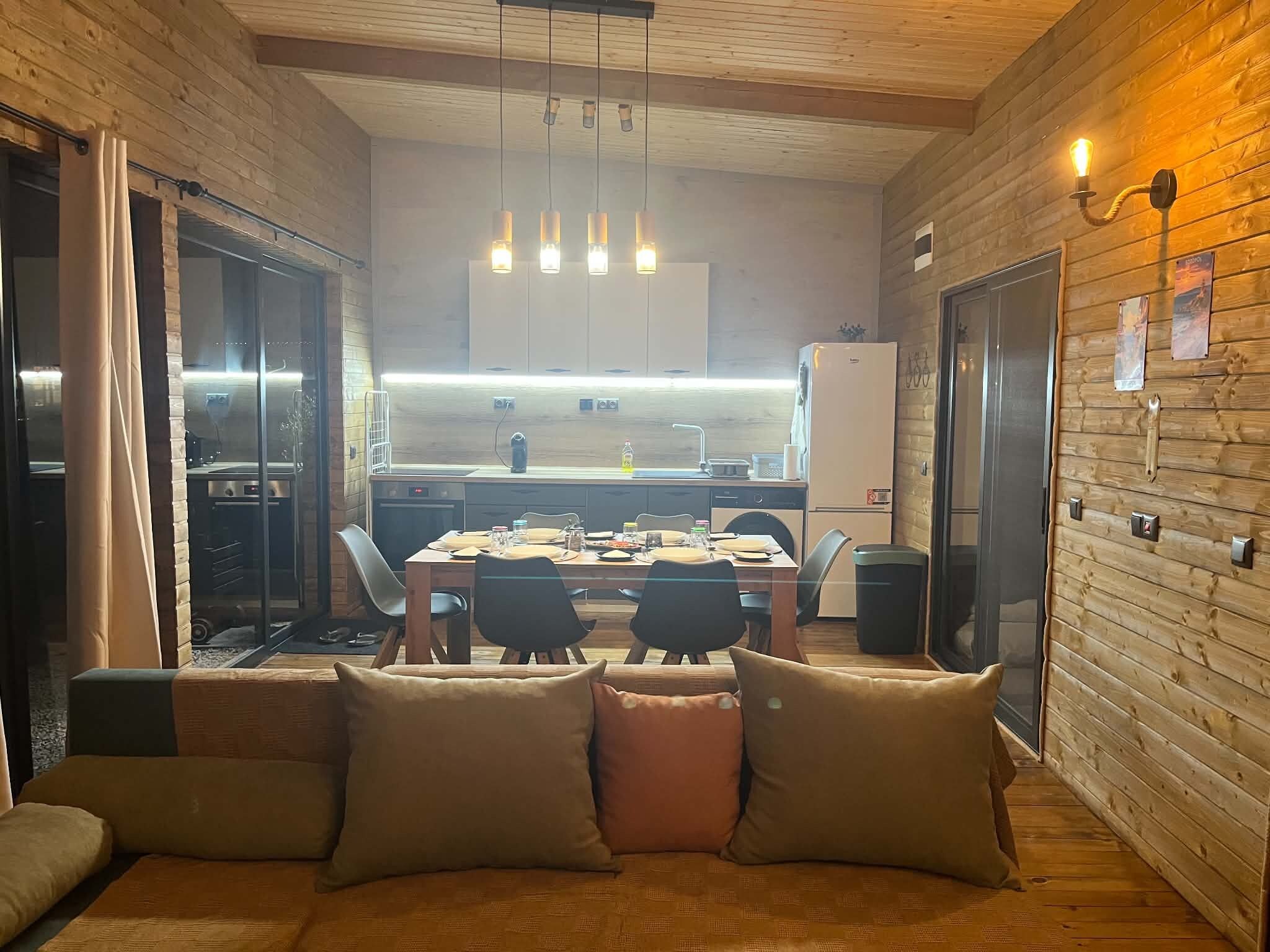 A cozy dining area with a wooden table and chairs, a sofa with cushions, and a kitchen in the background.