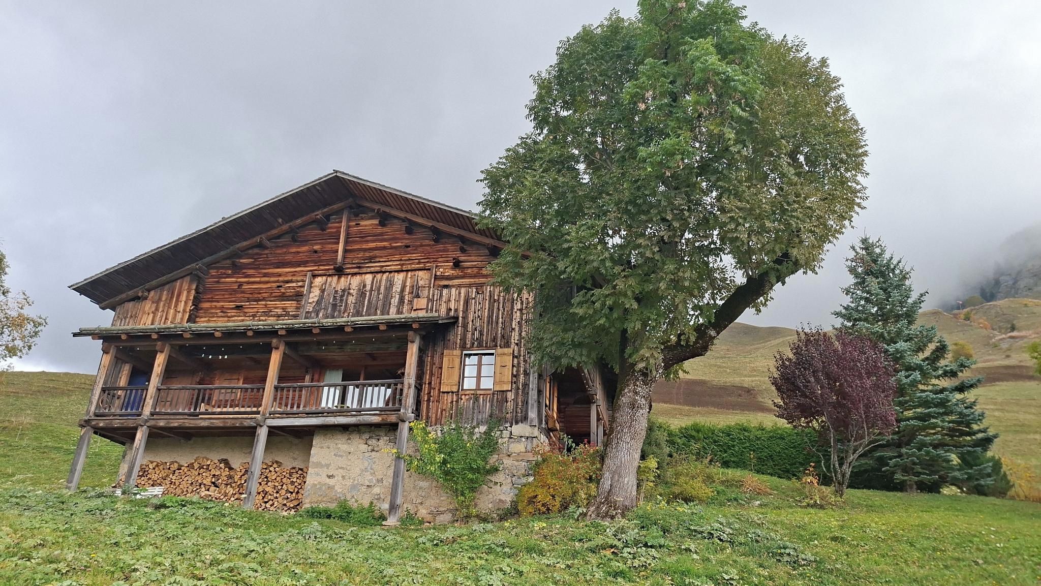Un chalet en bois rustique doté d’un balcon, entouré de verdure et avec une colline en arrière-plan.