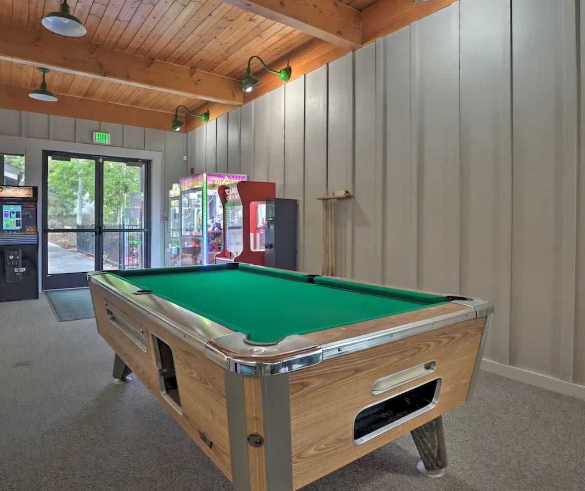 A pool table in a room with wooden ceiling and walls, a door leading outside, and a vending machine in the background.