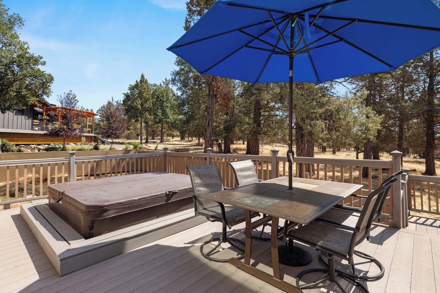 An outdoor deck with a table and chairs, a blue umbrella, and a view of a building and trees.
