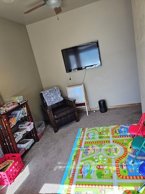 Una habitación con piso alfombrado, televisor montado en la pared, una silla, una pequeña mesa con una pizarra y un tapete de juegos colorido.