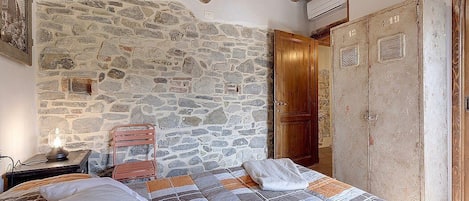 Un dormitorio con pared de piedra, vigas de madera en el techo, una cama con colcha estampada, una mesita de noche con lámpara y un armario de madera.