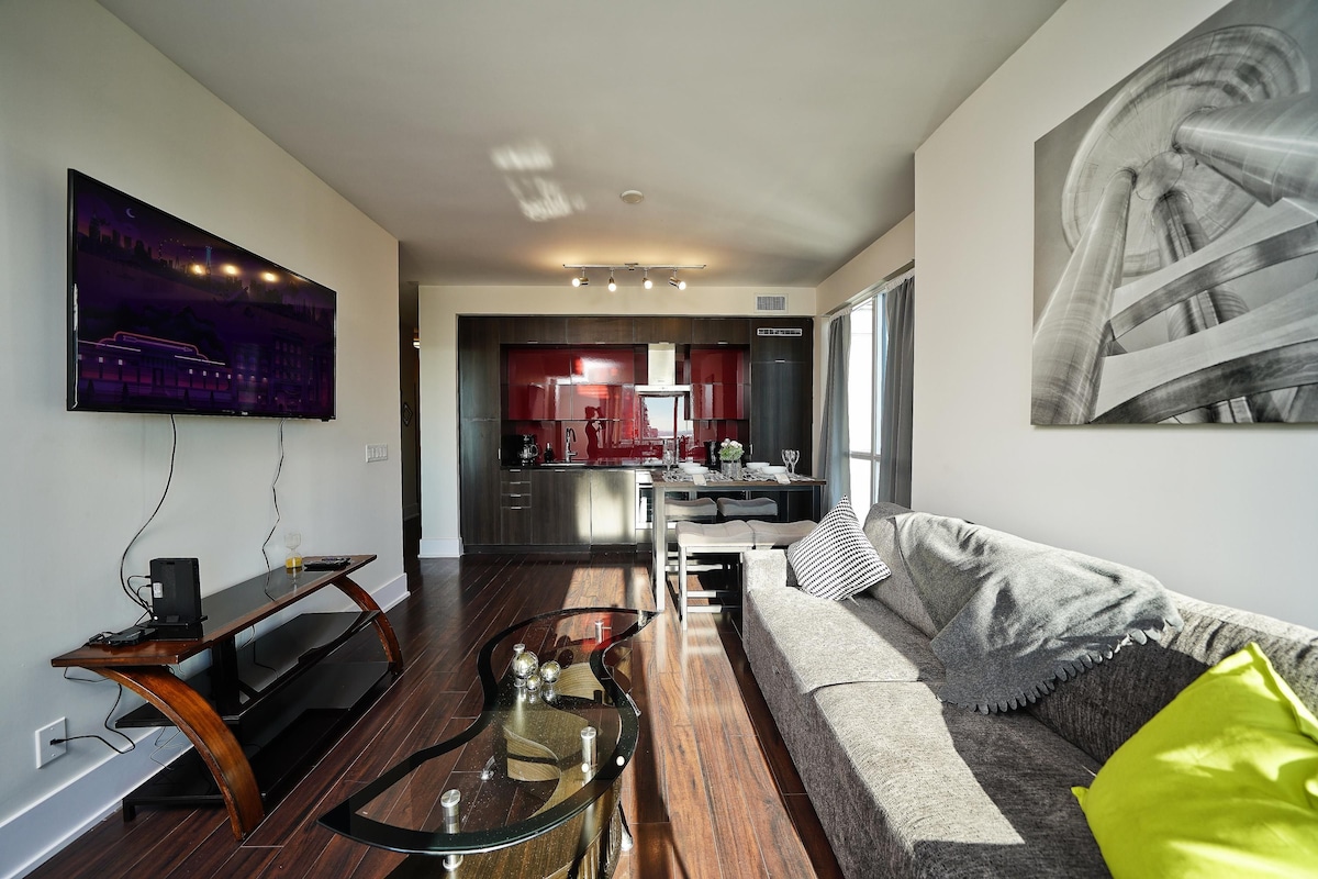 Una sala de estar moderna con un televisor de pantalla plana, una mesa de centro de vidrio, un sofá con cojines y una zona de comedor al fondo.