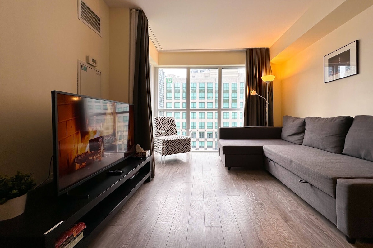 Una sala de estar moderna con un televisor de pantalla plana, un sofá gris y un ventanal con vistas a la ciudad.