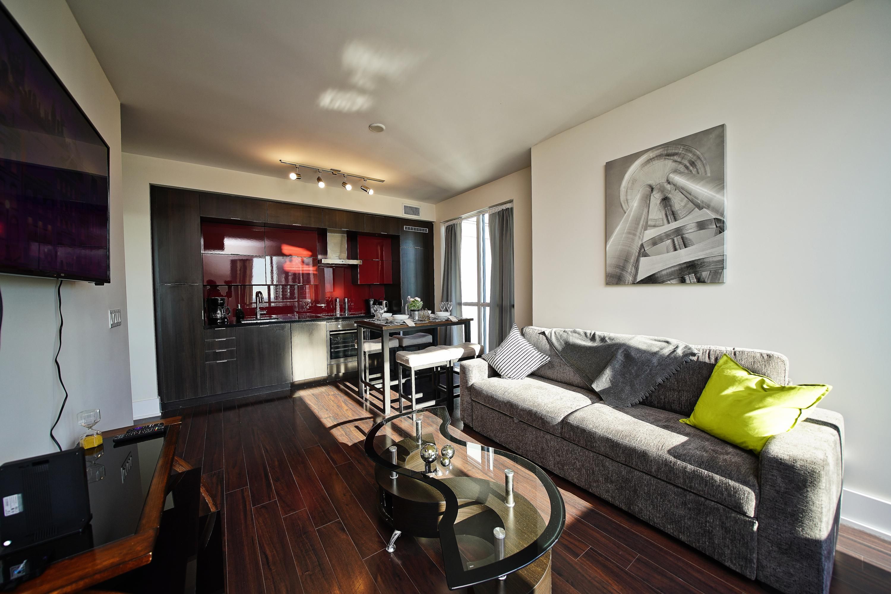 A modern living room with a glass coffee table, a sofa with a green pillow, a kitchen in the background, and a mounted artwork.