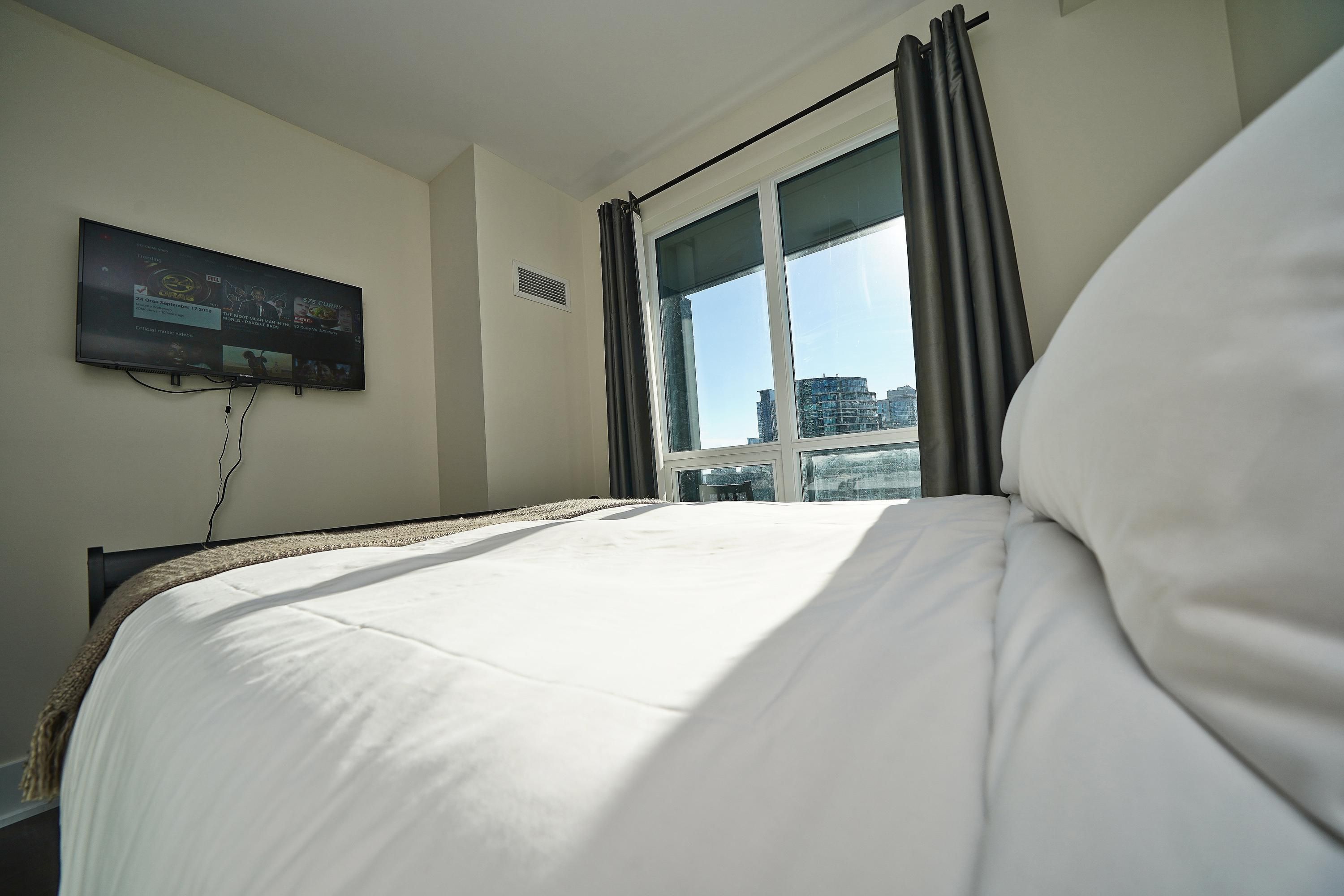 A hotel room with a bed, a TV mounted on the wall, a window with curtains, and a view of buildings outside.