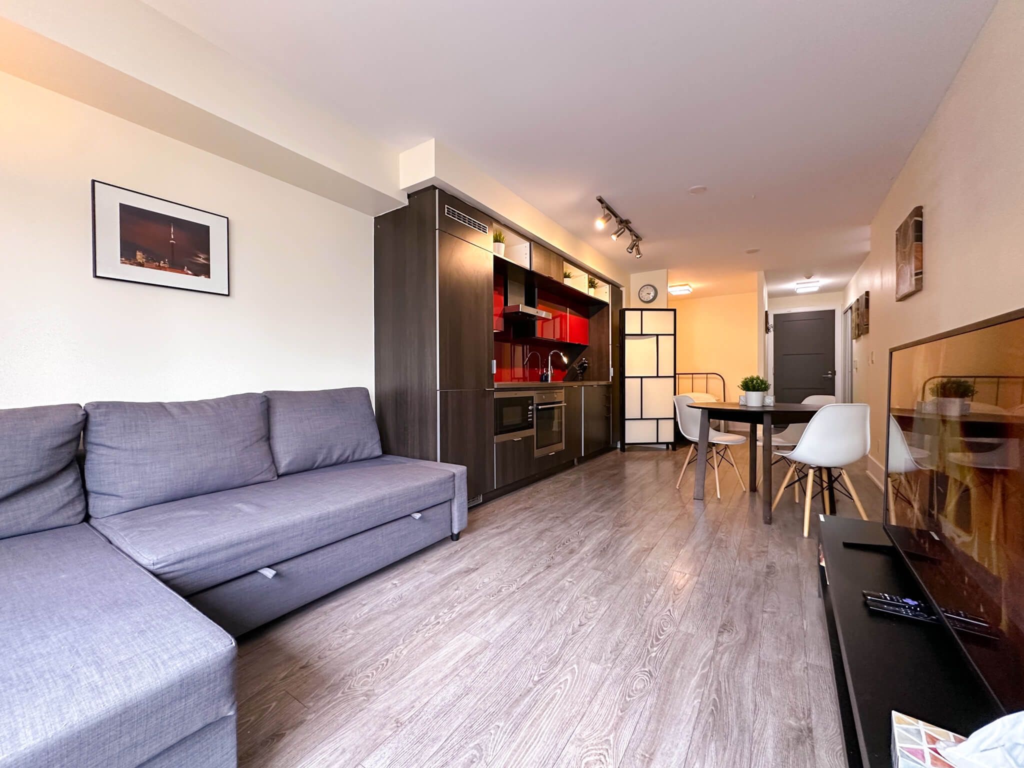 A modern living space with a grey sofa, wooden flooring, a kitchen with red cabinets, and a dining area with white chairs.