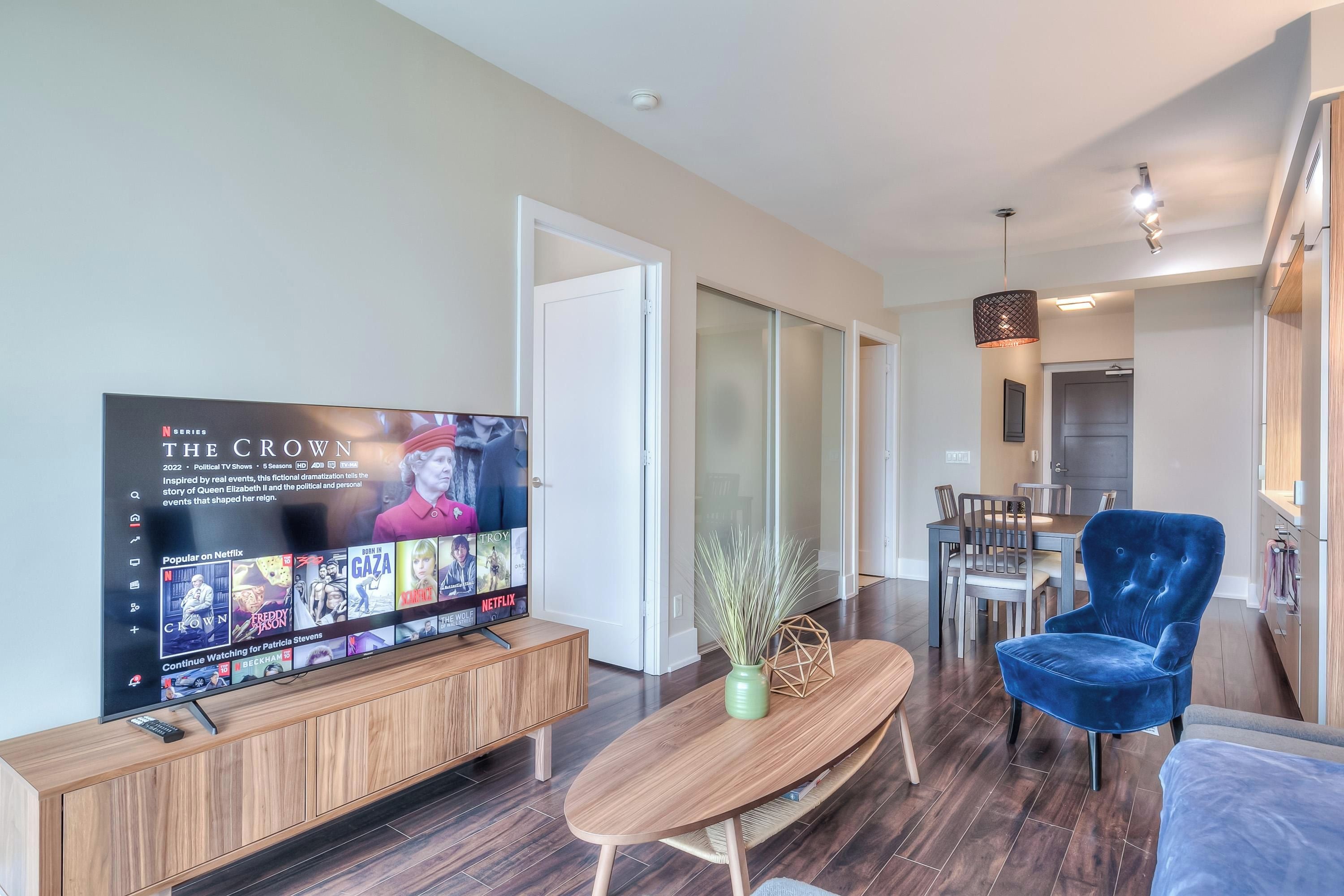 A modern living room with a flat-screen TV displaying 'The Crown' series, a wooden coffee table, a blue velvet armchair, and a dining area in the background.