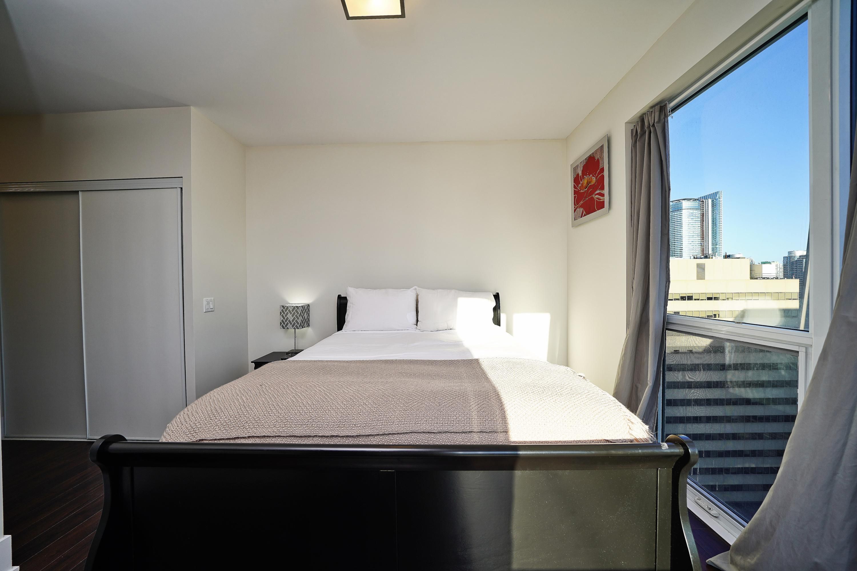 A hotel room with a bed, a window with a view of buildings, a door, and a wall-mounted light fixture.