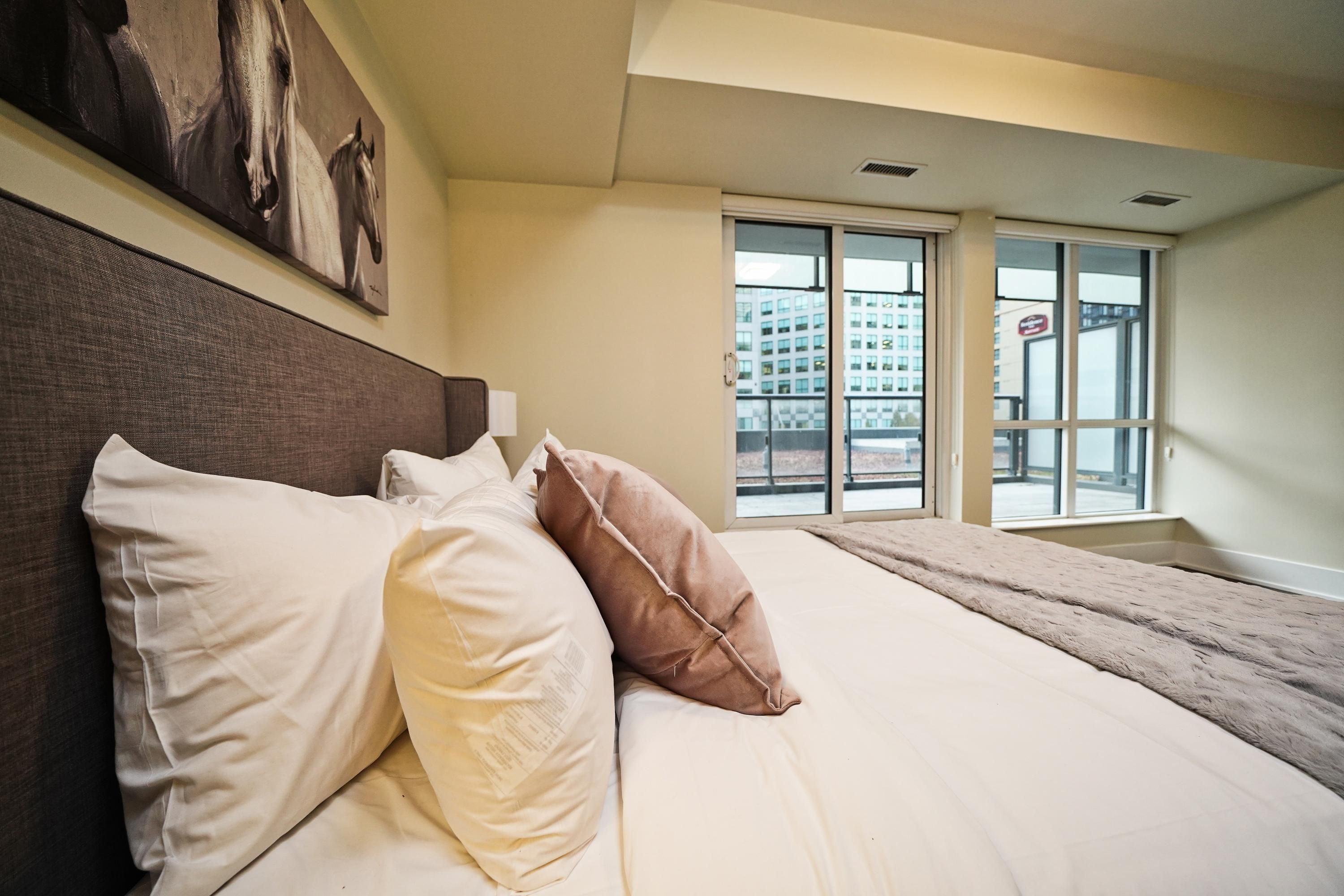 Een slaapkamer met een groot bed, kussens, een hoofdeinde, een raam met uitzicht op gebouwen en een schilderij aan de muur.