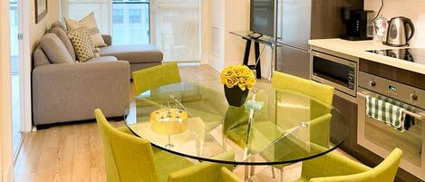 A modern kitchen with a dining area, featuring a glass table, green chairs, and a sofa with cushions.
