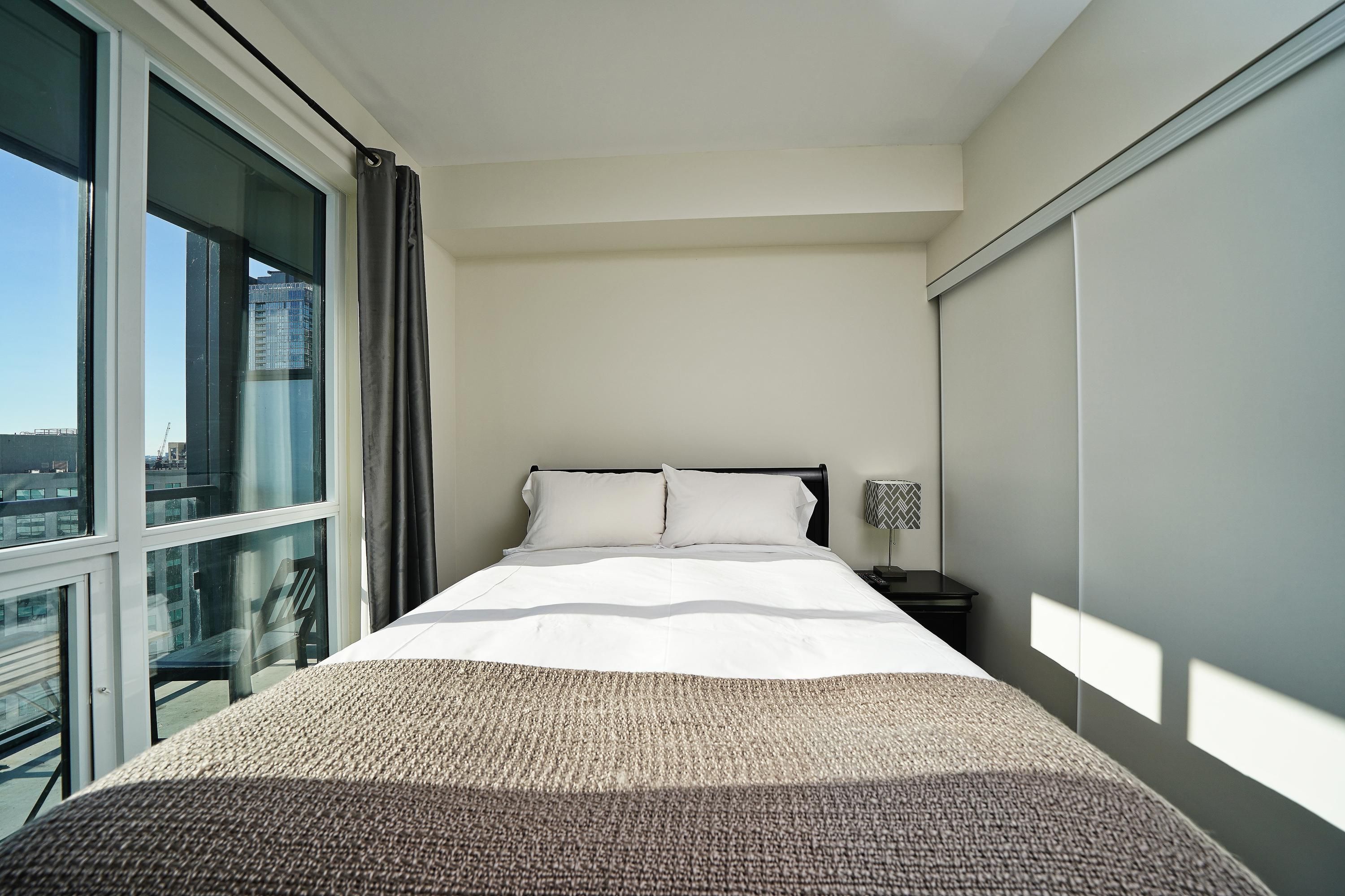 A hotel room with a large bed, a nightstand, a lamp, and a view of a cityscape through a large window.