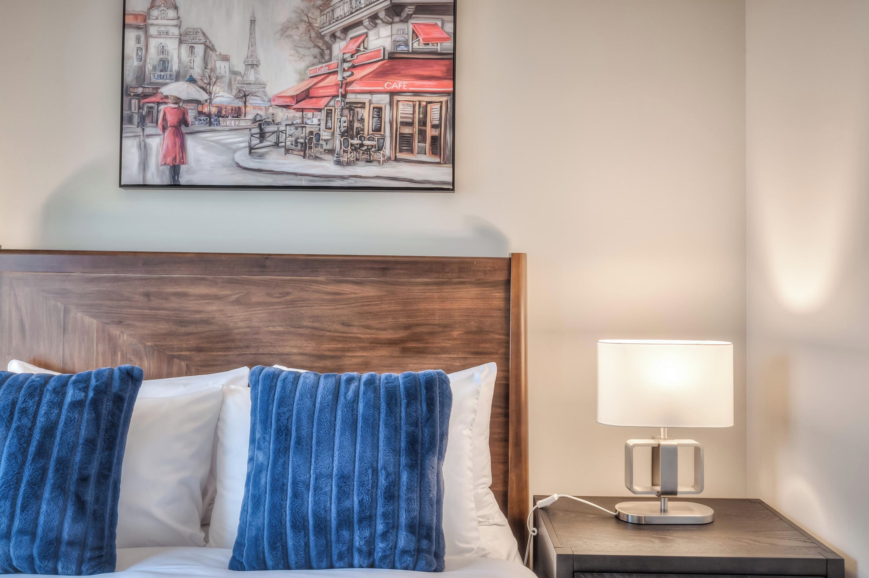 Habitación de hotel con cabecera de madera, cama con almohadas azules y blancas, mesita de noche con lámpara y un cuadro con una escena callejera en la pared.