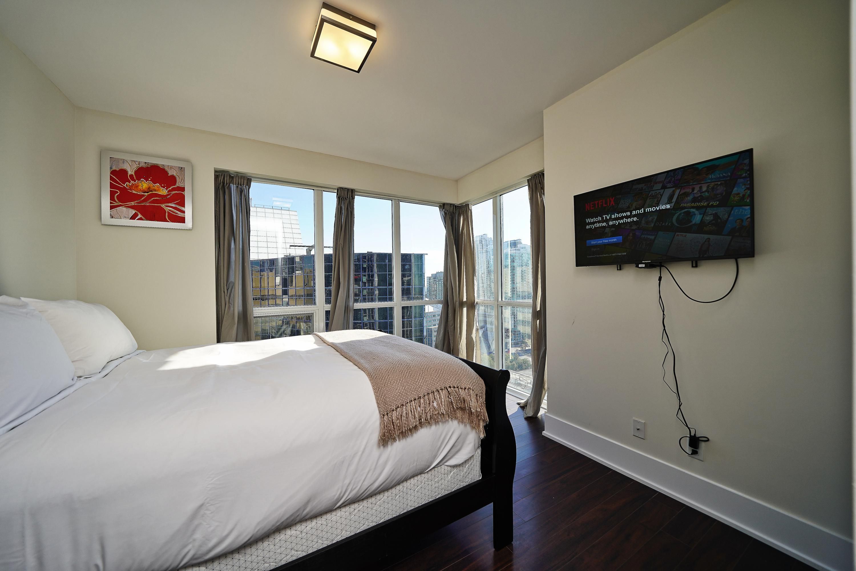 A bedroom with a large bed, a wall-mounted TV, and a window with curtains.