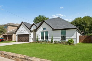 A two-story house with a garage, a well-maintained lawn, and a wooden fence. - DFW retreat with pool, hot tub, and back yard oasis (Irving)