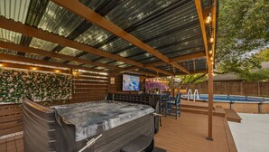 A hot tub with a wooden deck, a pool, and a covered patio area with a mural and string lights. - DFW retreat with pool, hot tub, and back yard oasis (Irving)
