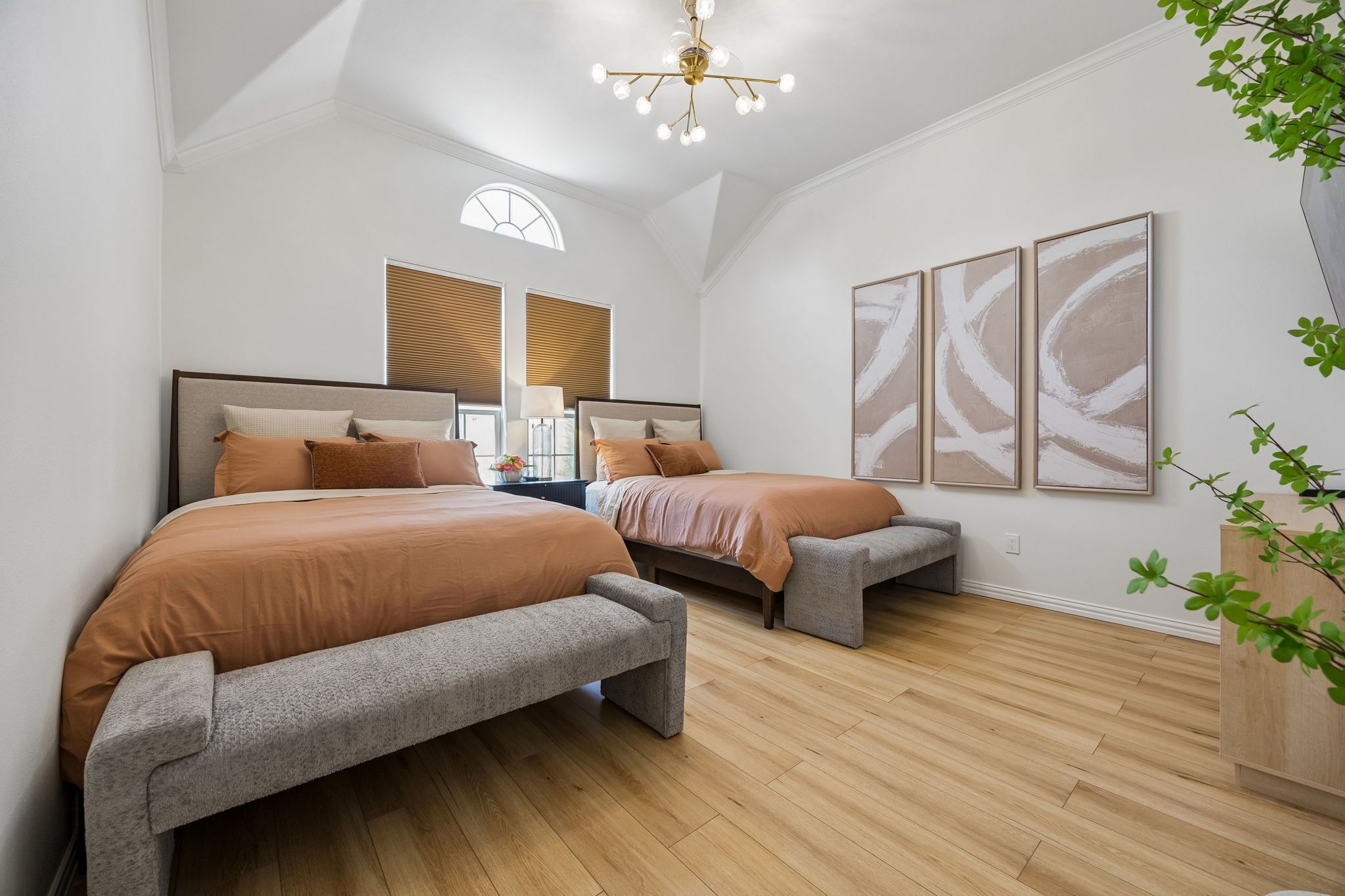 A bedroom with two beds, wooden flooring, a ceiling light fixture, and wall-mounted artwork.