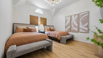 A bedroom with two beds, wooden flooring, a ceiling light fixture, and wall-mounted artwork.
