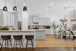 Een moderne keuken met witte kasten, een centraal eiland met barkrukken, en een eetgedeelte met een kroonluchter.