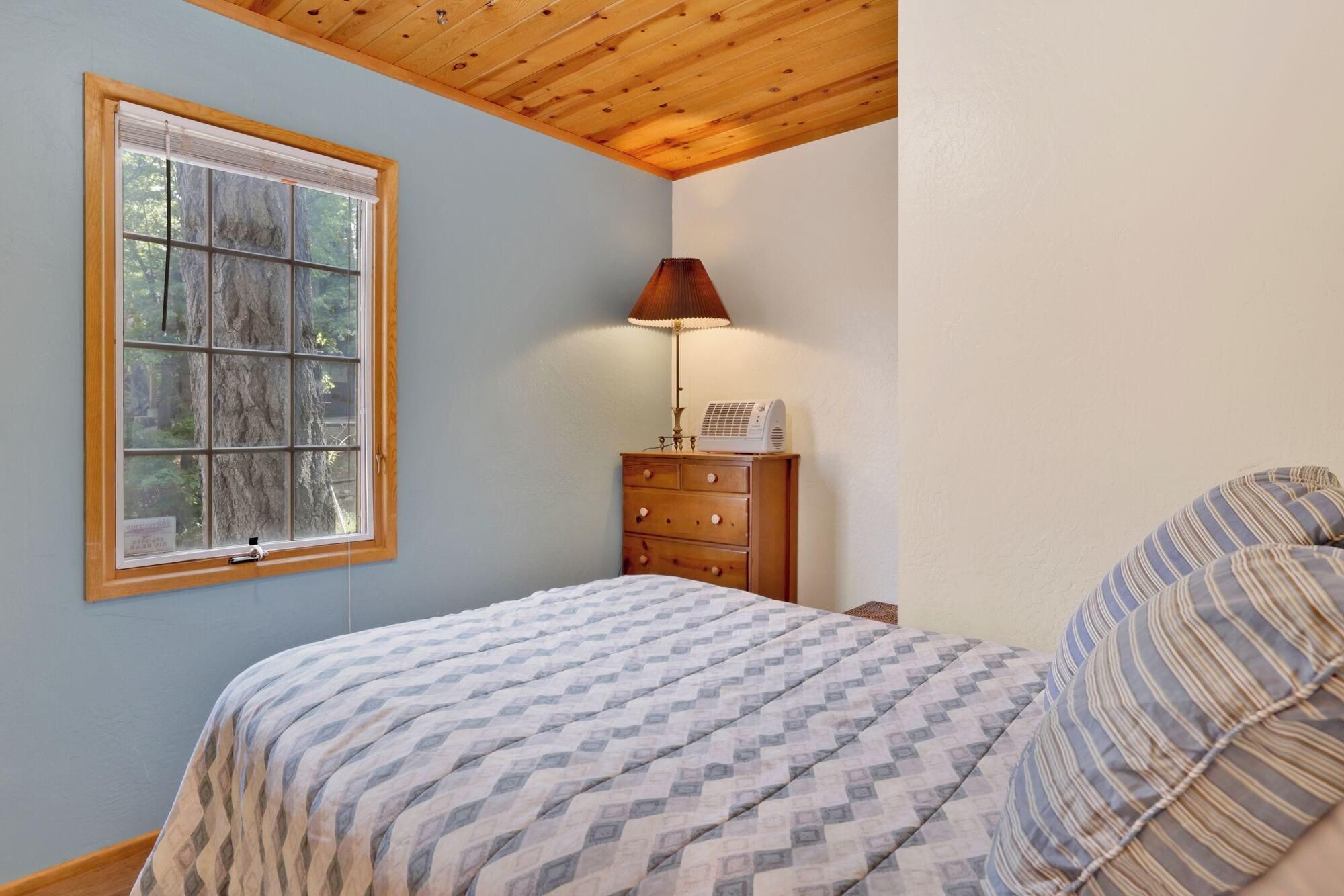 Een slaapkamer met een bed, een dressoir, een lamp en een raam met uitzicht op bomen.