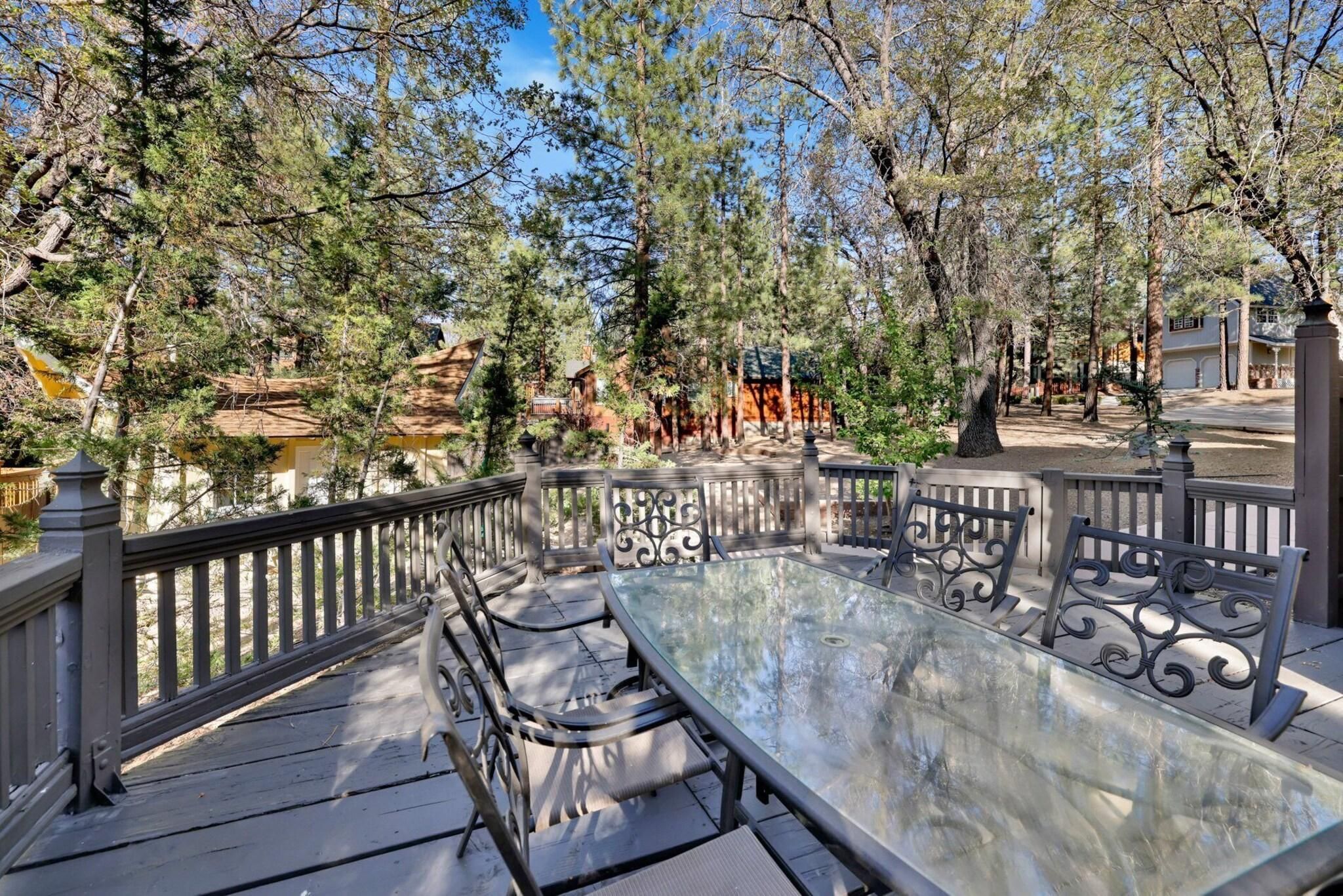 An outdoor dining area with a glass table and metal chairs, surrounded by trees and a fence.