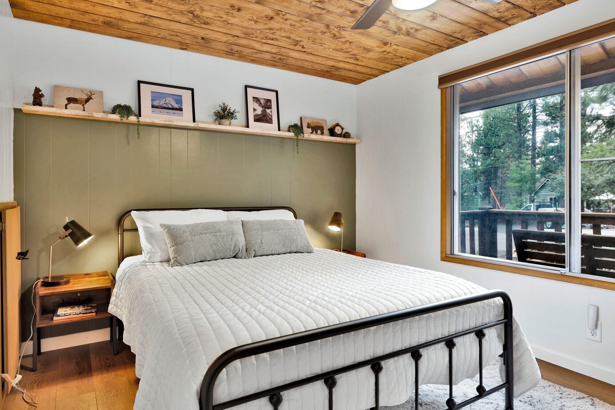 A bedroom with a bed, bedside lamps, a wooden headboard, a window with a view, and a shelf with pictures and small items.