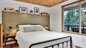 A bedroom with a bed, bedside lamps, a wooden headboard, a window with a view, and a shelf with pictures and small items.
